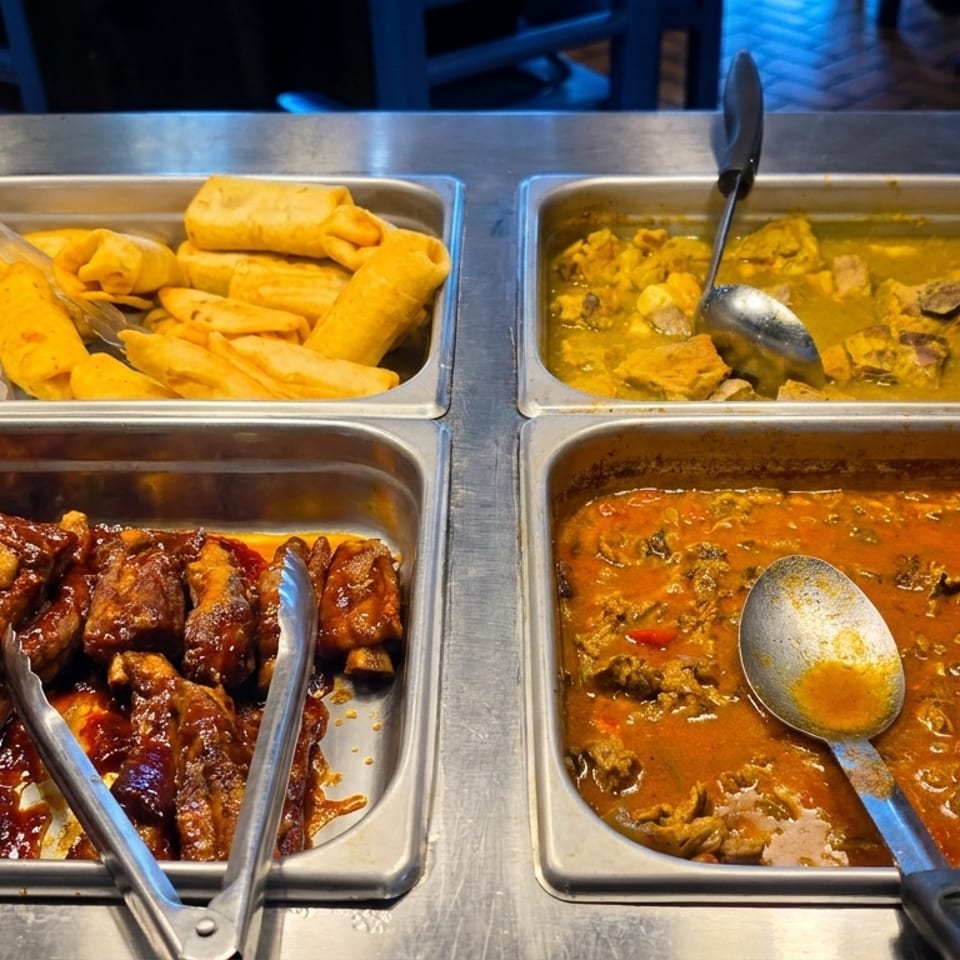 Four buffet trays filled with barbecue ribs, yellow plantains, chicken curry, and a red stew.